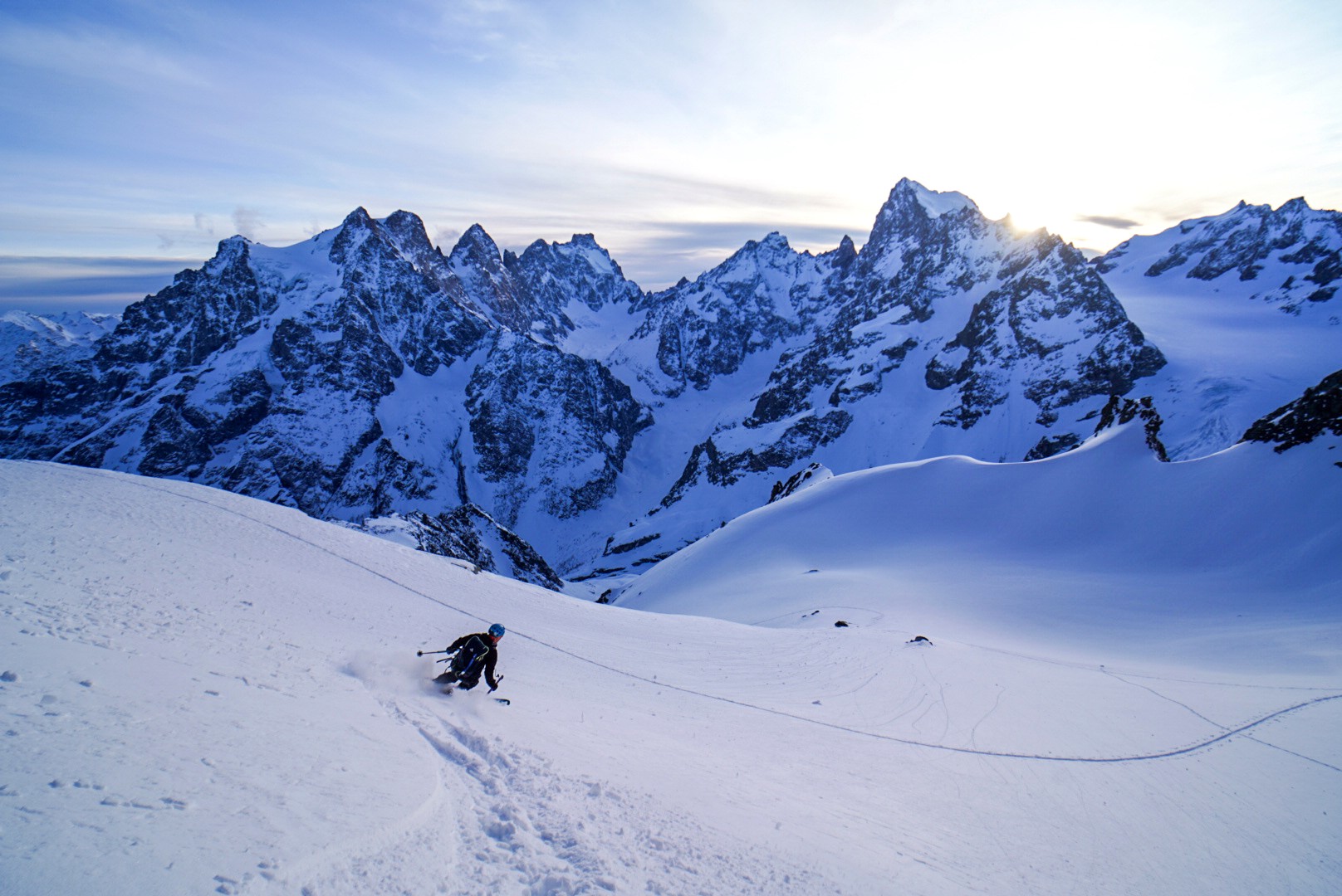 Skieur isolé dans une pente de neige, face à une chaîne de sommets rocheux éclairés par le soleil, Tour des Agneaux (Écrins)