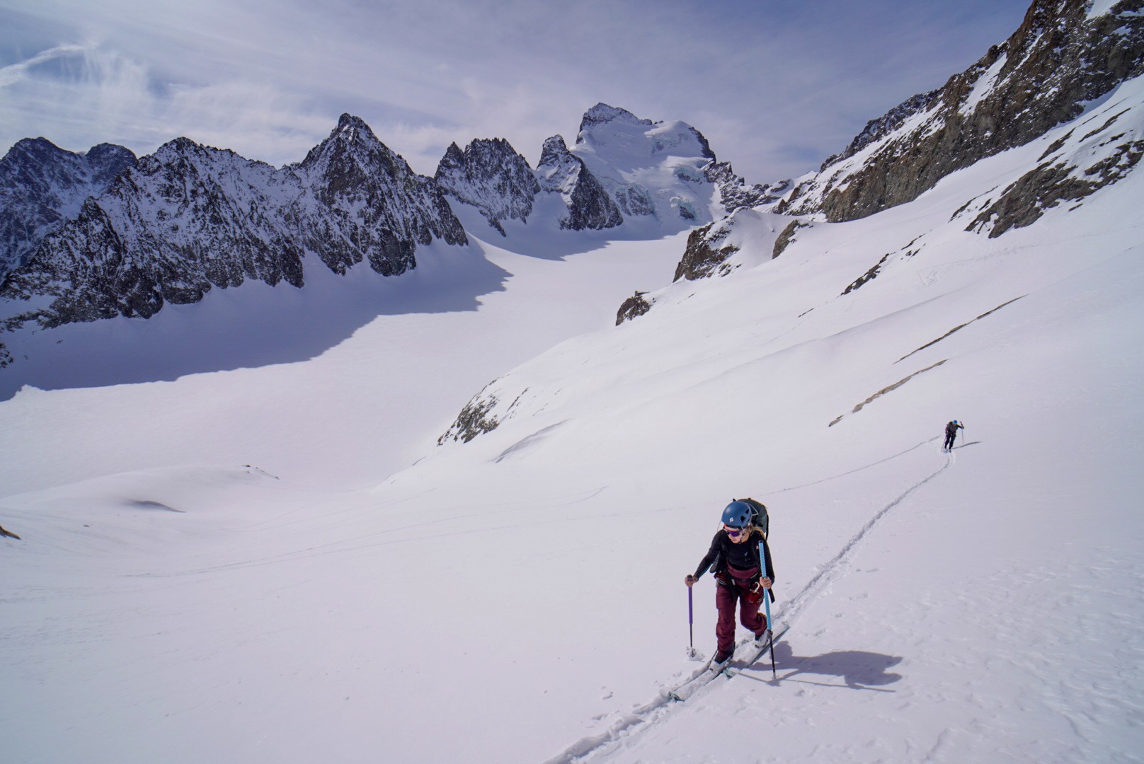 Skieuse en montée sur un glacier face à une crête rocheuse, un autre skieur plus haut, Tour des Agneaux (Écrins)