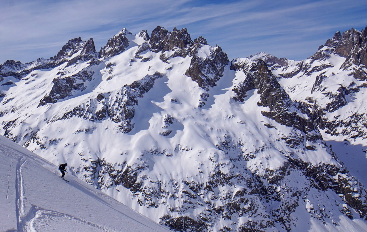 Skieur en descente sur une pente, face à une grande paroi rocheuse enneigée, Tour des Agneaux (Écrins)