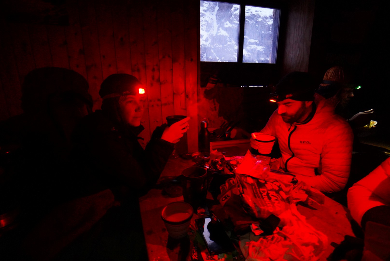 Trois personnes attablées dans un refuge, éclairées par la lumière rouge de frontales, Tour des Agneaux (Écrins)