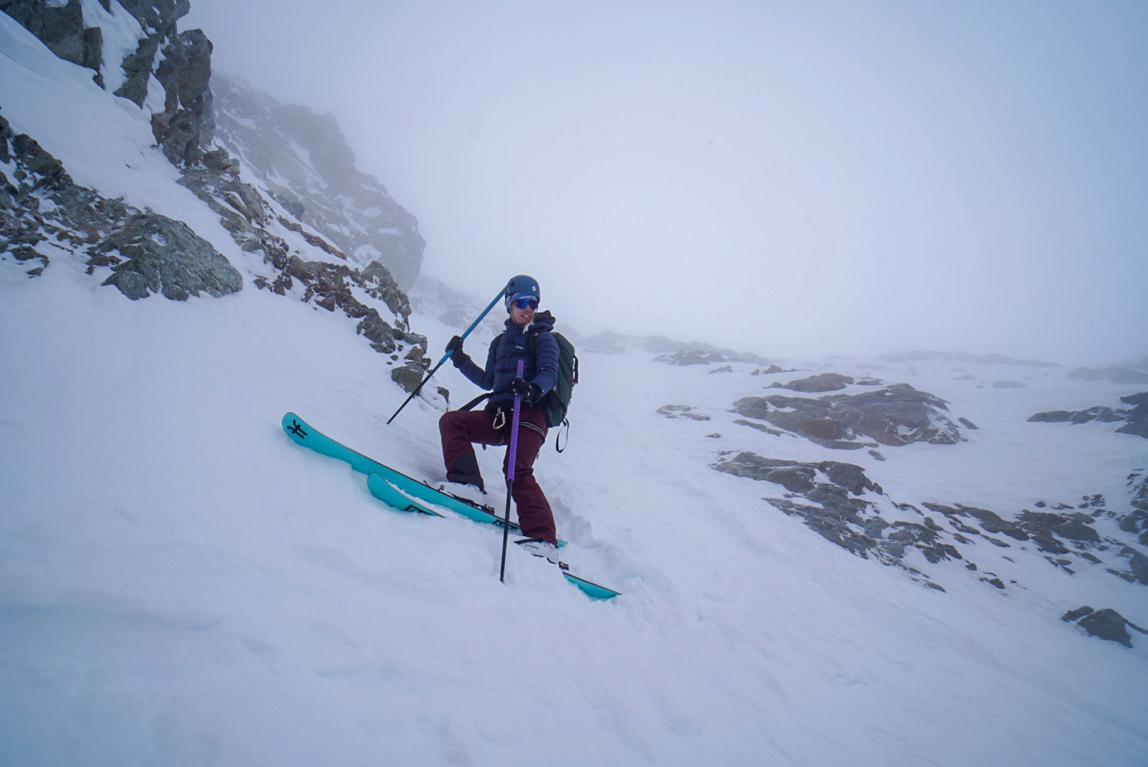 Skieuse assise dans une pente raide, skis sur la neige, au pied d'une barre rocheuse dans la brume, Tour des Agneaux (Écrins)
