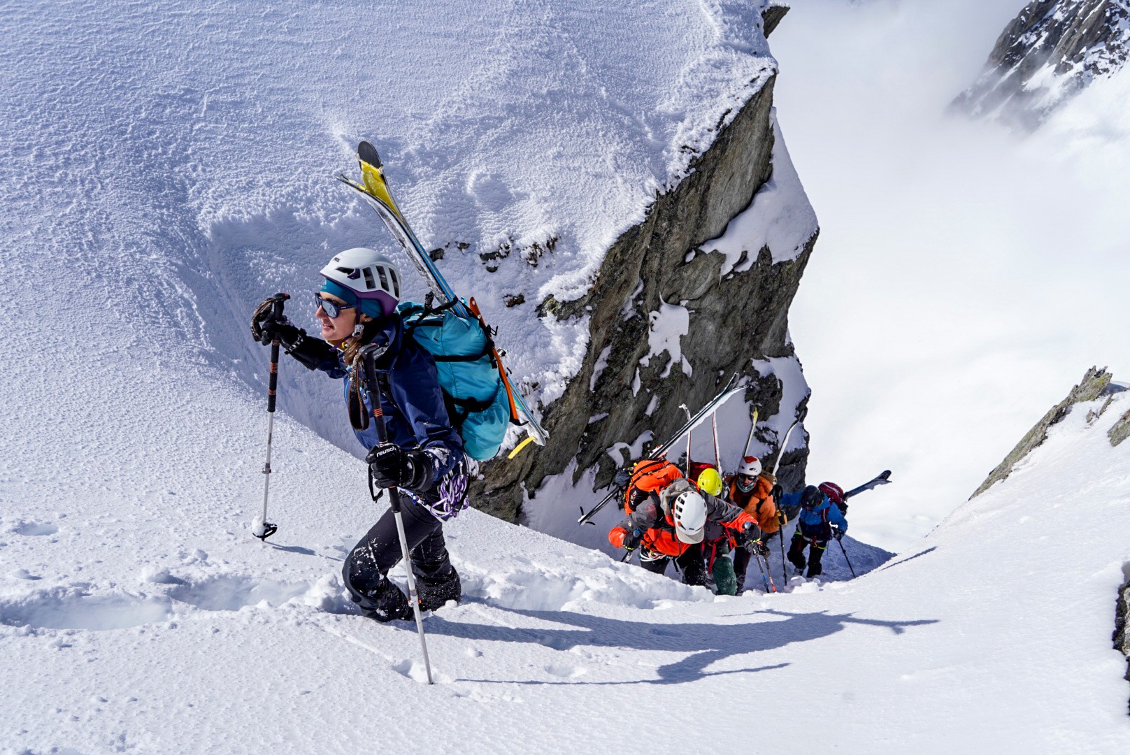 Montée à pied dans un couloir de neige étroit bordé de rochers, skis sur le sac, cordée en contrebas