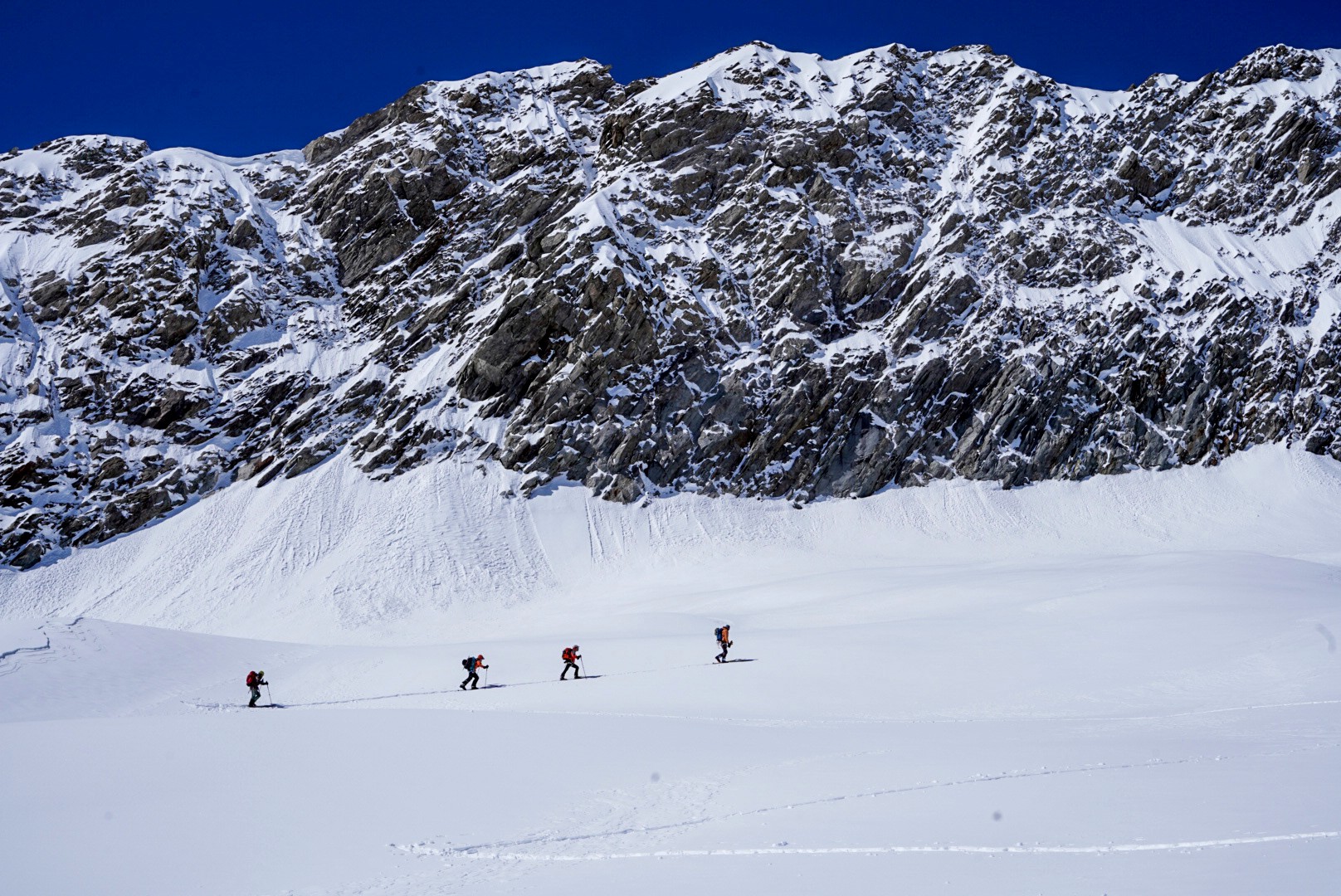 Quatre skieurs de randonnée traversant un vaste glacier sous un mur rocheux, Dammastock