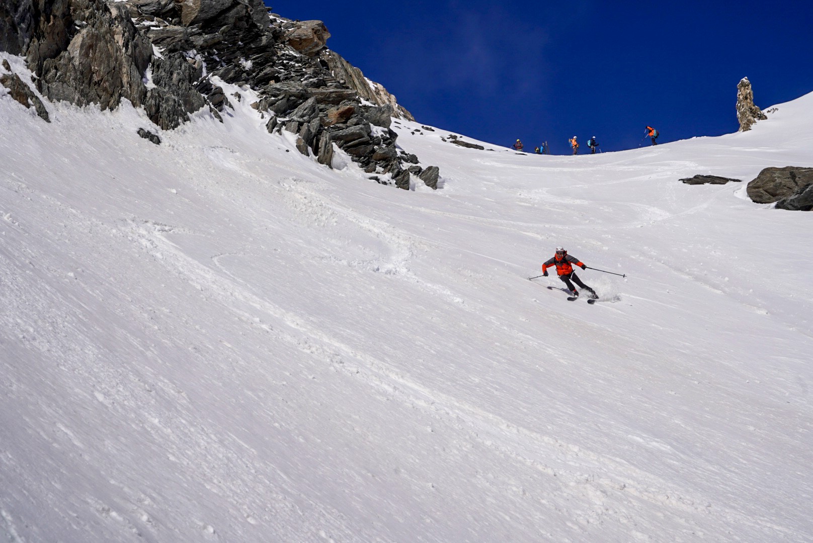 Skieur descendant une pente de neige vers un col rocheux, le reste du groupe en haut, Dammastock