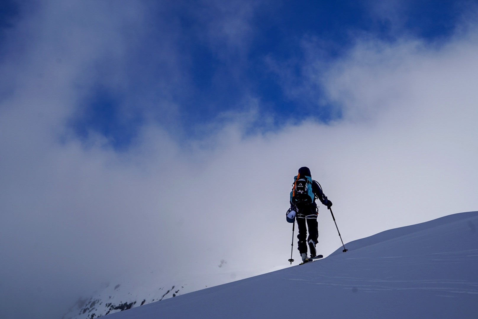Skieur de randonnée en montée sur une crête enneigée, entre nappes de nuages et éclaircies bleues, traversée du Dammastock (Suisse)