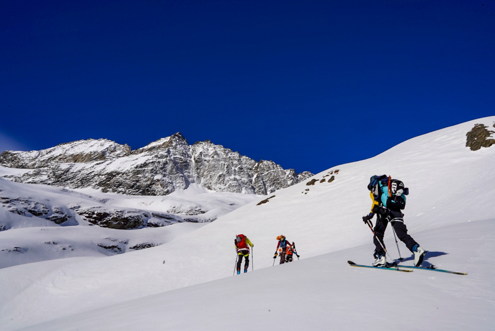 Trois skieurs de randonnée en montée dans un vallon enneigé, sommet rocheux en arrière-plan
